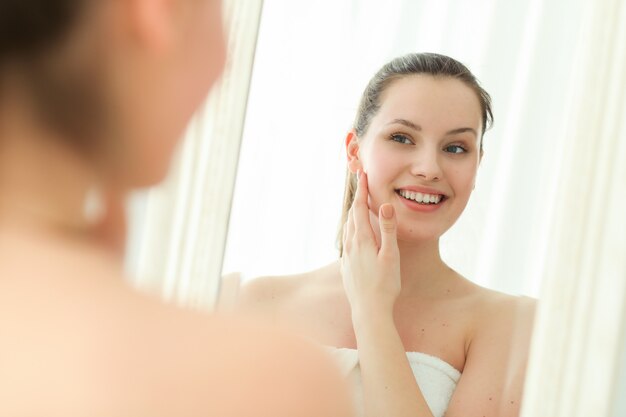 Woman with towel on body after shower, looking in mirror　美肌　韓国コスメ　人気